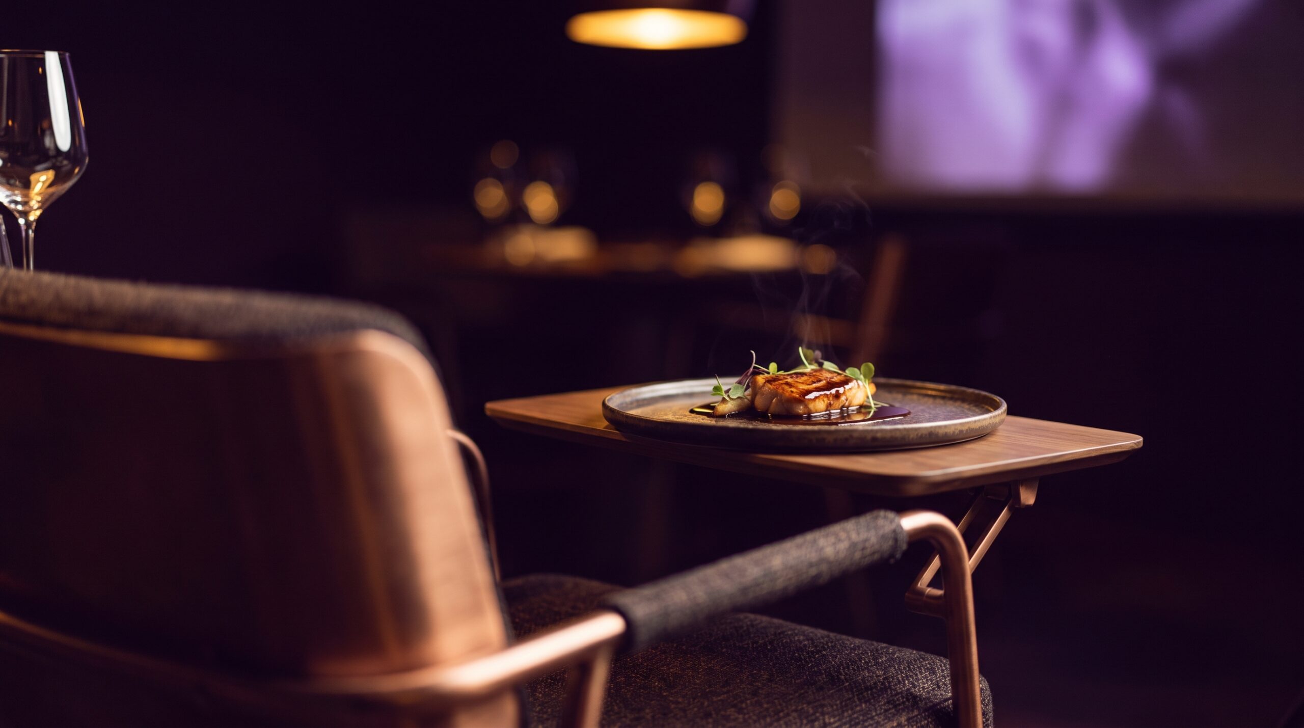 A single fine-dining course on a dark ceramic plate, resting on the fold-out wooden table of a high-backed lounge chair in a darkened dining room lit by one warm overhead lamp, with a wine glass in the foreground.