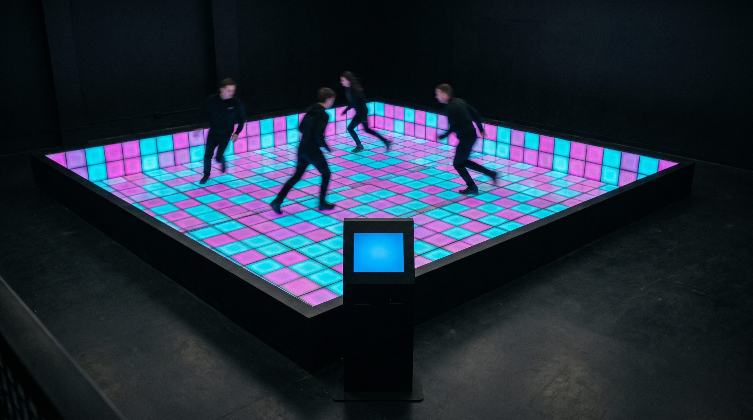 Four people run across a square arena floor of glowing cyan and magenta light-up tiles in a dark room, seen from above, with a plain black self-service kiosk standing unattended at the front edge.