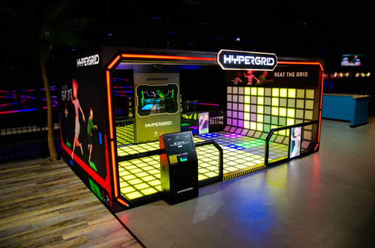 The HYPER GRID interactive LED floor attraction seen at an angle in a dark venue beside a go-kart track, its floor and wall tiles lit yellow and green, with an unattended self-service kiosk at the front edge.