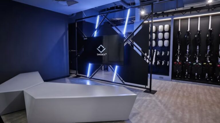 A Sandbox VR venue lobby with a lit Sandbox VR screen and a wall of haptic vests and headsets. Photo courtesy of Sandbox VR.